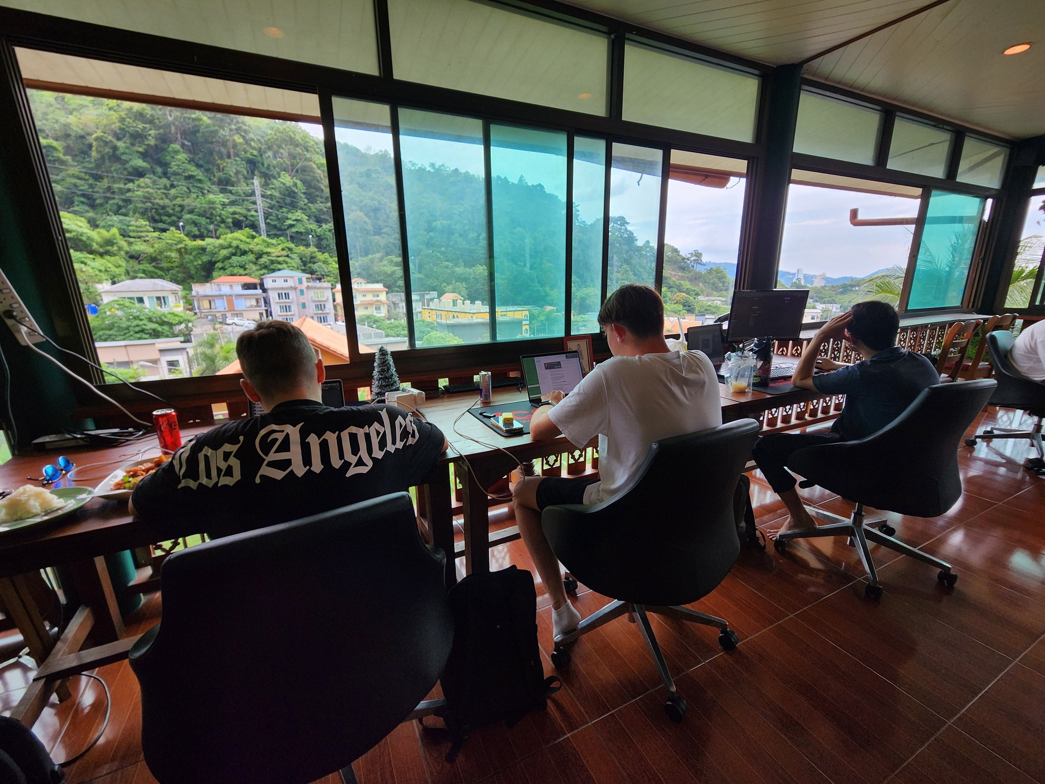 Three people working on laptops at a shared desk facing windows with a hillside village view at Denz CoWorking, Patong Phuket