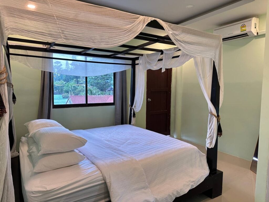 Four-poster canopy bed with white bedding beside a window with green hillside view at Denz WorkStay, Patong Phuket