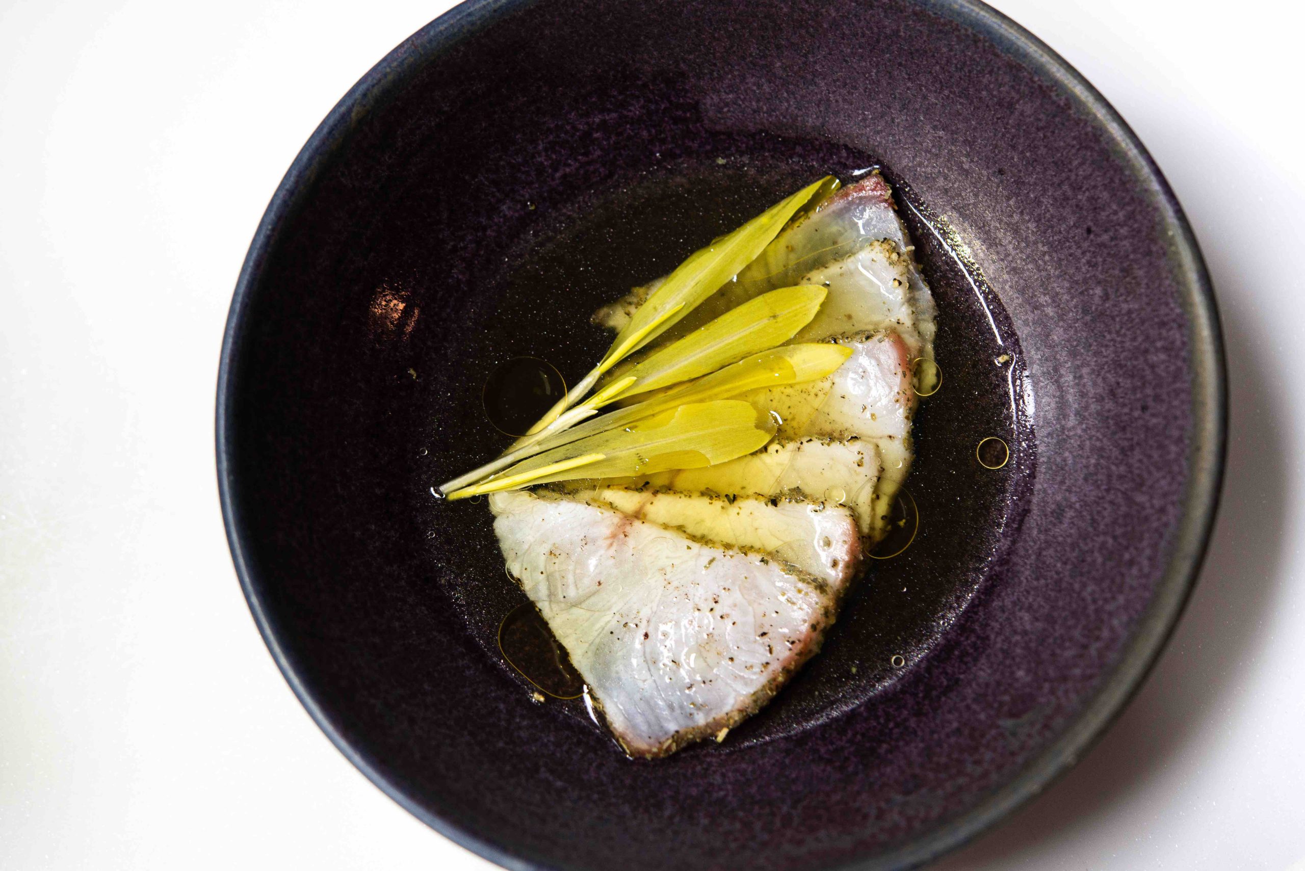 Cured fish crudo with yellow petals in oil in a dark bowl at Heh contemporary restaurant, Old Town Phuket
