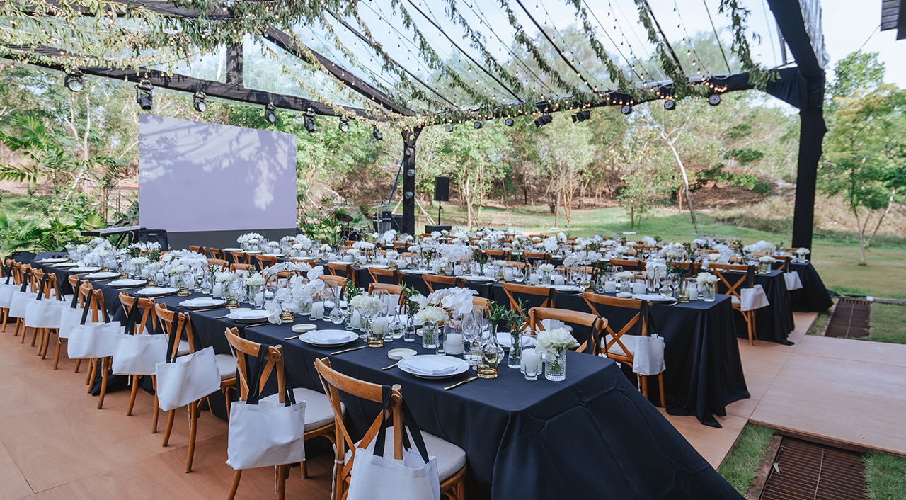 Long tables with black linens, white florals and crossback chairs under a clear tent at Jampa restaurant in Bang Tao, Phuket