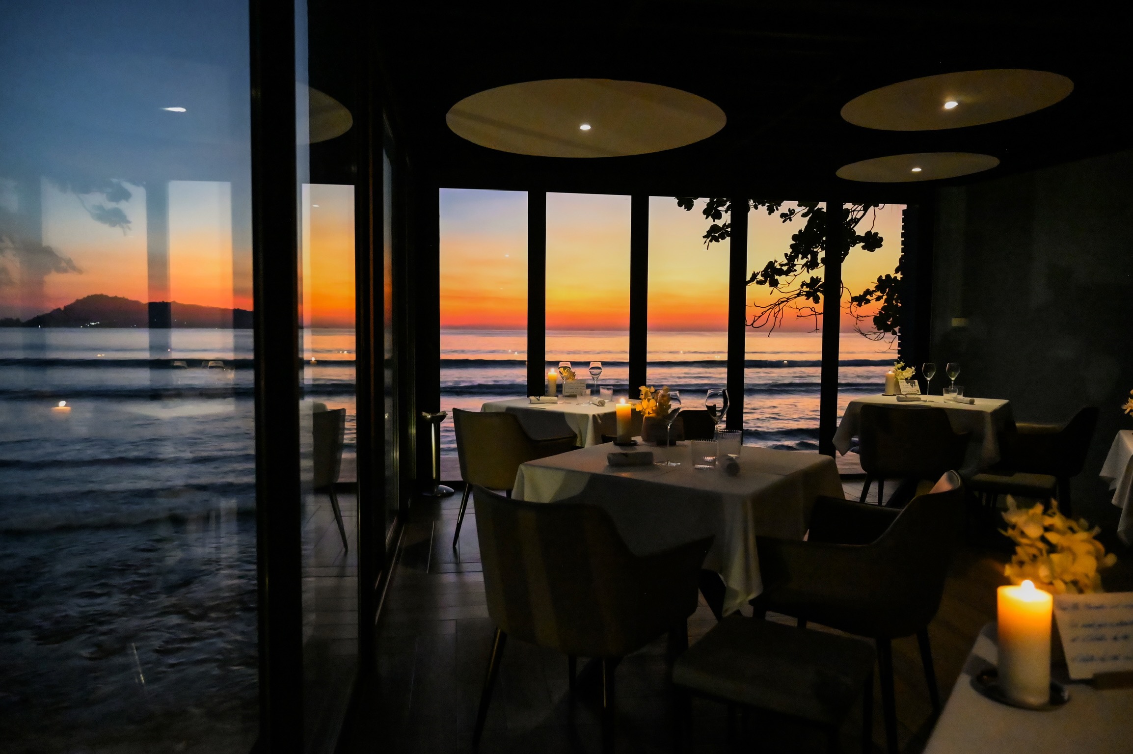 Candlelit white-clothed tables with a sunset sea view inside L'Arôme by the Sea restaurant in Patong, Phuket