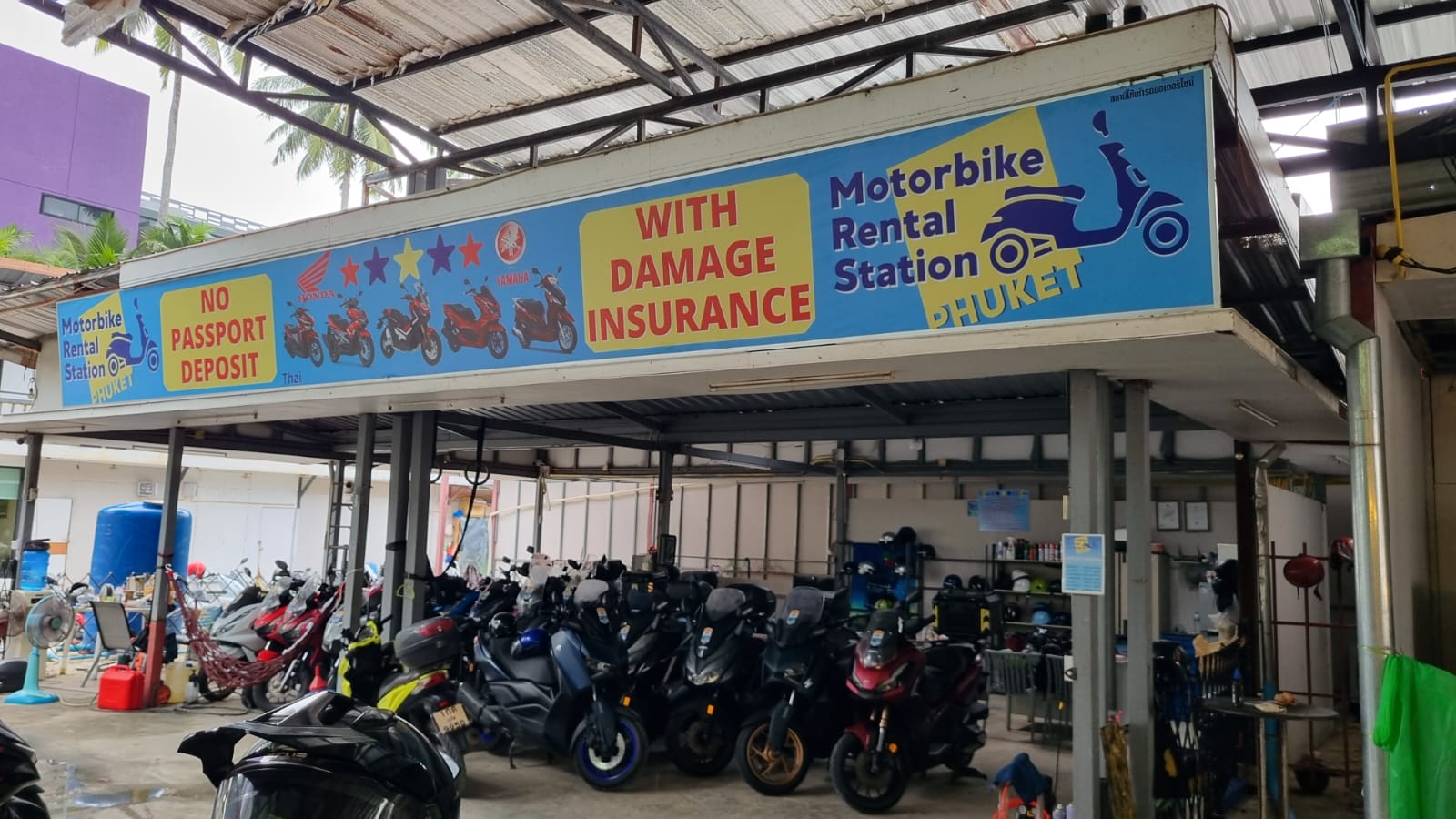Covered garage with parked scooters under signs for no passport deposit and damage insurance at Motorbike Rental Station Phuket, Patong