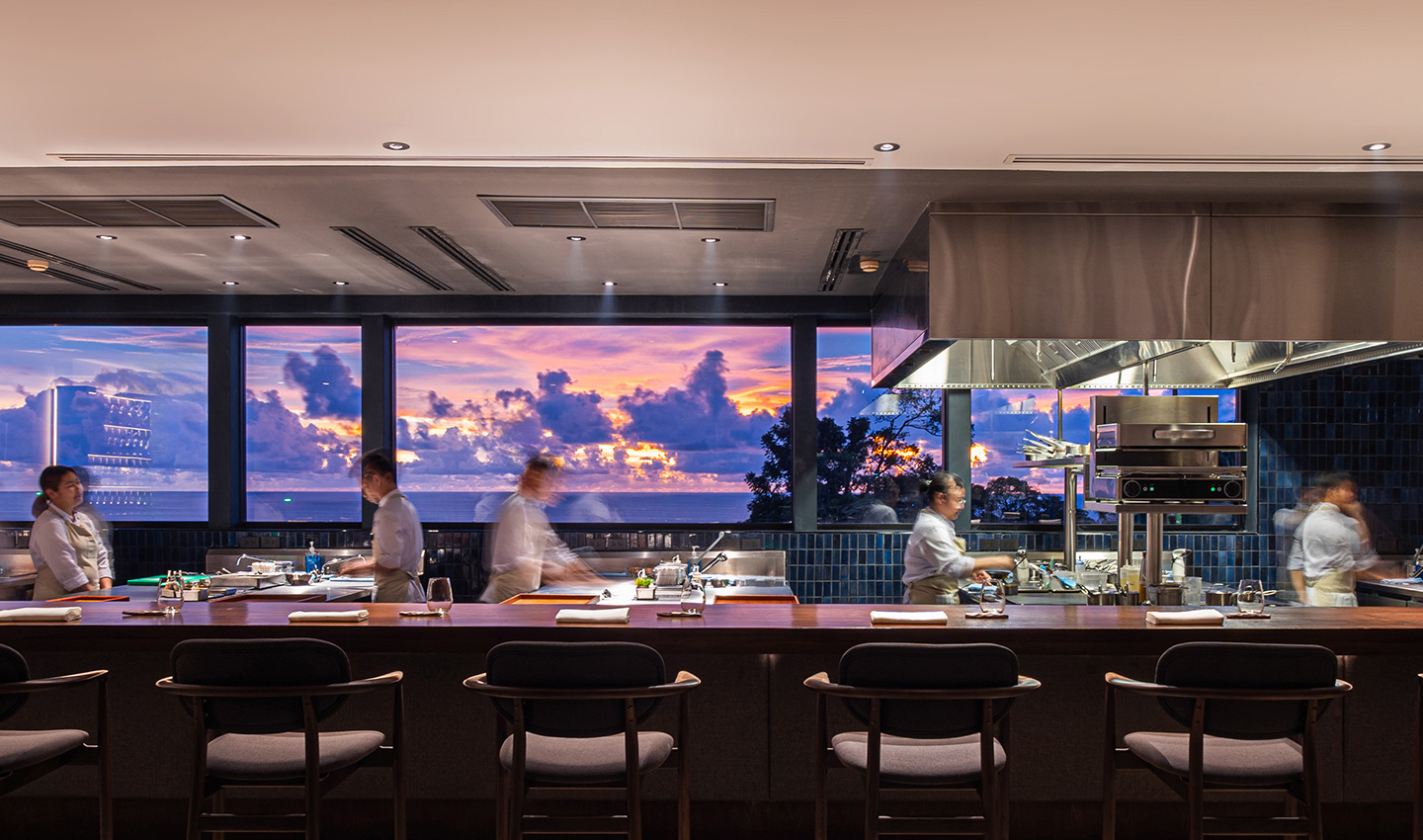 Chefs at work behind the open-kitchen chef's counter with a sunset sky through the windows at PRU in Bang Tao, Phuket.