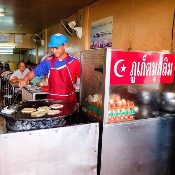 Cook frying rounds of roti on a large griddle beside stacked eggs and a red Halal sign at Roti Thaew Nam in Phuket Old Town, Phuket.