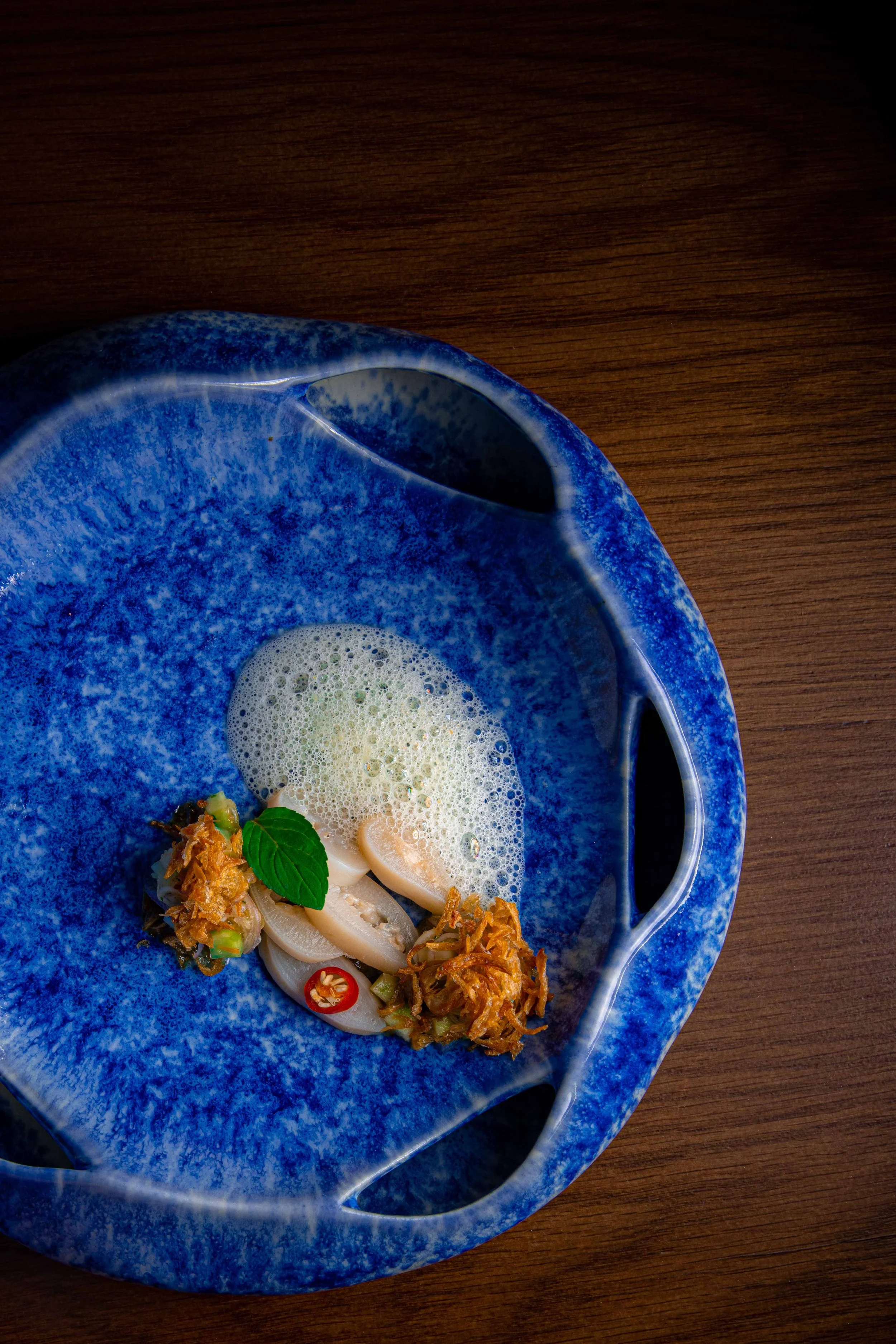 Plated dish of sliced seafood with crispy shallots, chilli, mint and foam on a blue glazed bowl at Samut, a Southern Thai restaurant in Rawai, Phuket.