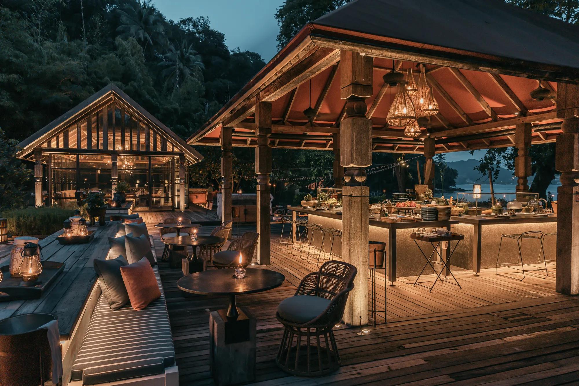 Lantern-lit wooden dining deck and thatched pavilion bar at dusk, Ta Khai seafood restaurant at Rosewood Phuket in Patong
