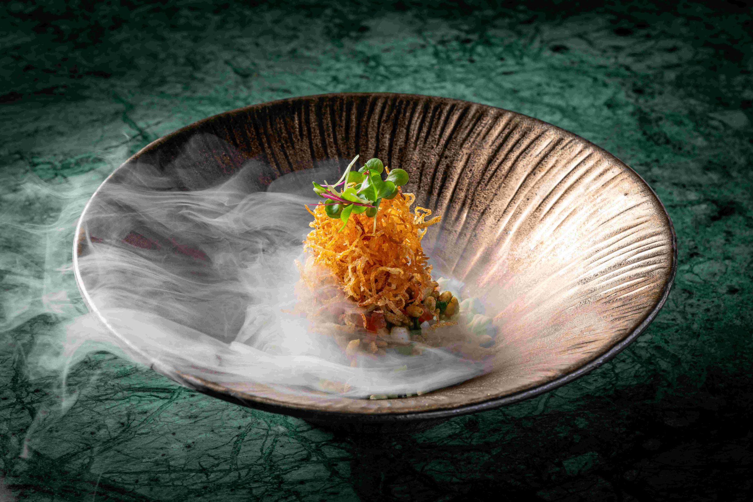 Dish topped with crispy fried garnish and microgreens amid dry-ice smoke in a bronze bowl at Tambu, an Indian restaurant in Patong, Phuket