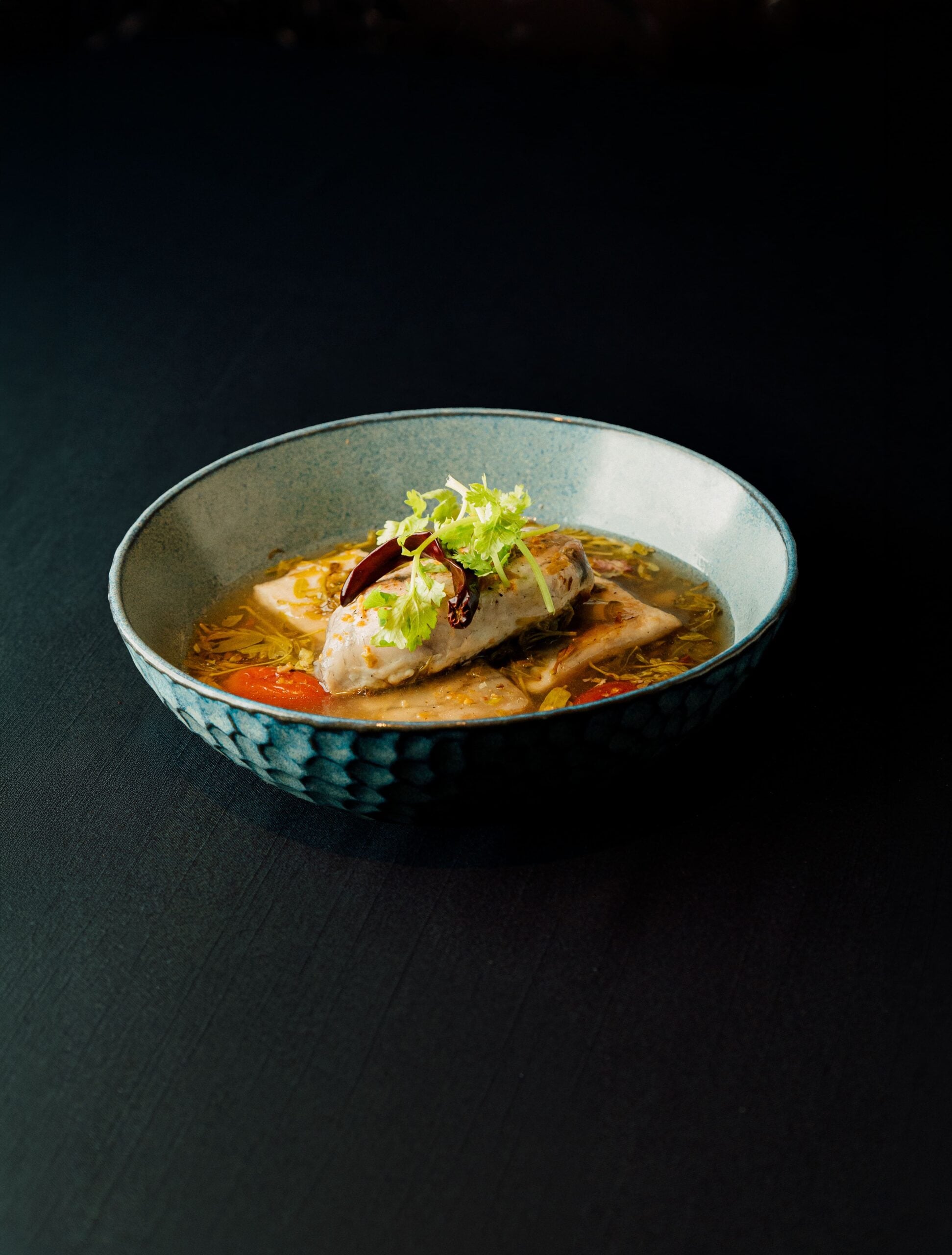 Fish fillet in a fragrant herb broth with tomato and chili in a blue ceramic bowl at Thai Library, a Thai restaurant in Bang Tao, Phuket
